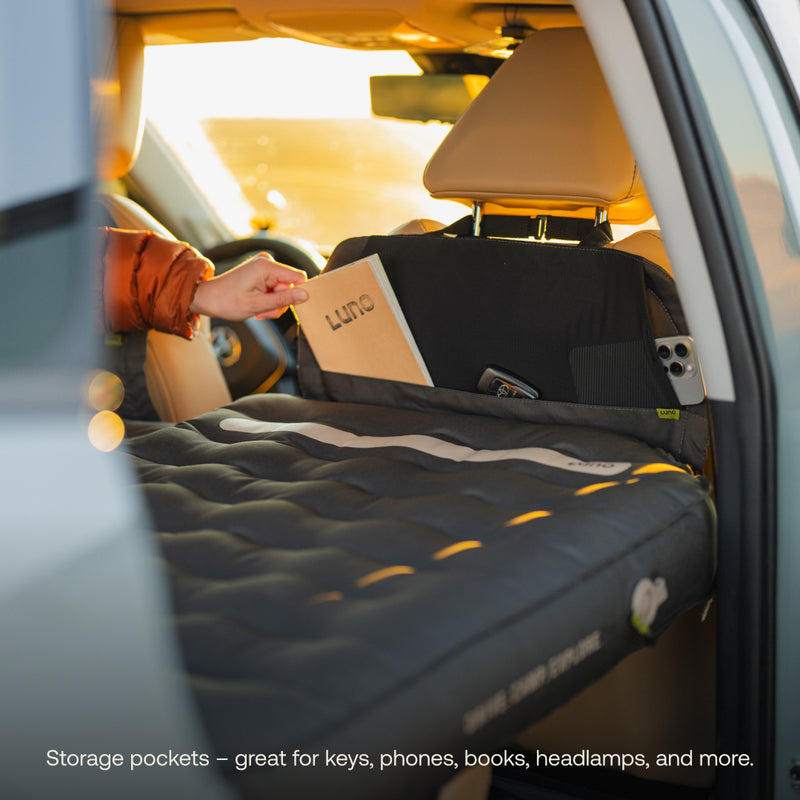 A person slides a book into the backseat storage pocket of a car equipped with the Luno® AIR Pro Vehicle Mattress. Sunlight highlights the cozy setup and ideal pockets for storing keys, phones, headlamps, and more.