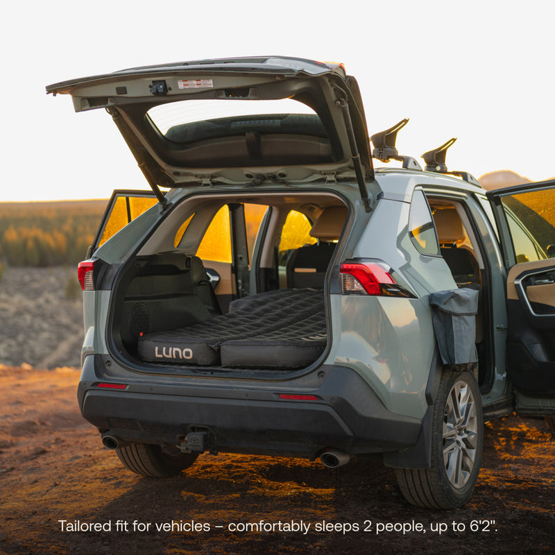 The open hatchback of a parked SUV shows a Luno® AIR Pro Vehicle Mattress, designed for two people up to 6'2". With the rear seats folded down, it forms a cozy sleeping area as the setting sun casts a warm glow over the scene.