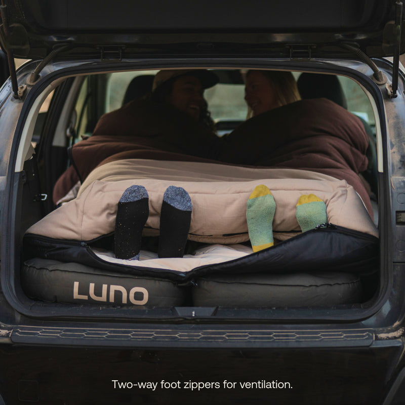 Two people lie in a car on a Luno air mattress under blankets. Their sock-clad feet peek out. Text: "Two-way foot zippers for ventilation." Ideal for car camping, the Luno® Fitted Sleeping Bag Comforter offers a double-wide fit and cushioned hood.