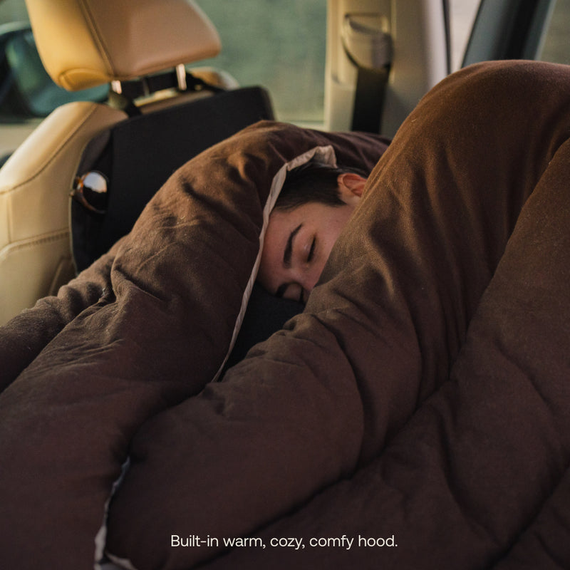 Inside a car's backseat, someone relaxes in a Fitted Sleeping Bag Comforter by Luno®, featuring a cozy brown design and cushioned hood. The text reads, "Built-in warm, cozy, comfy hood.
