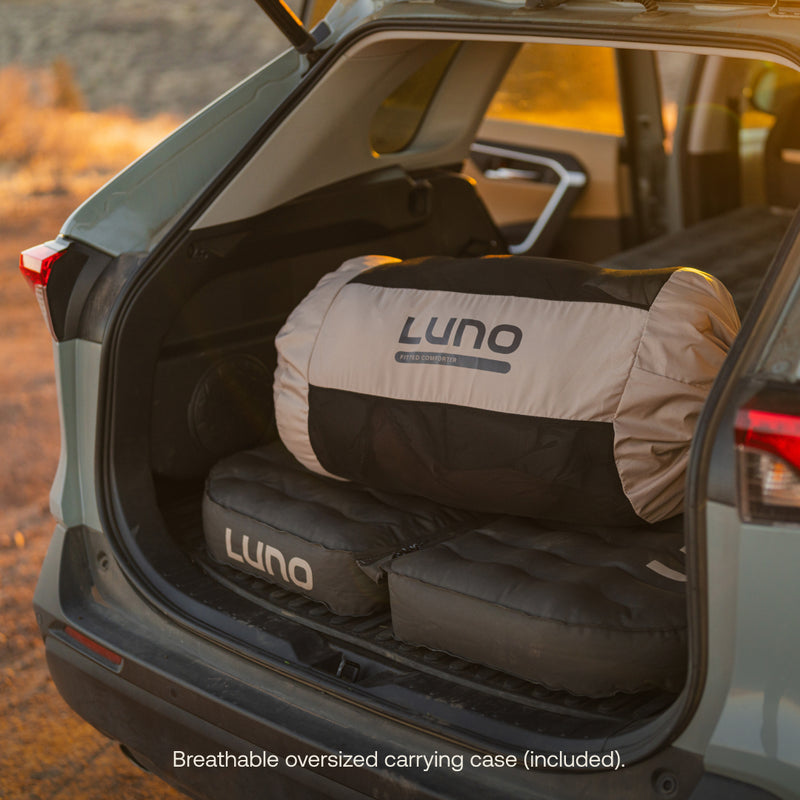 The image shows a car trunk with a Luno® inflatable mattress and an oversized carrying case for the Fitted Sleeping Bag Comforter, ideal for vehicle sleepers seeking comfort. Warm sunlight enhances the scene, highlighting the breathable case included for easy storage of the comforter.