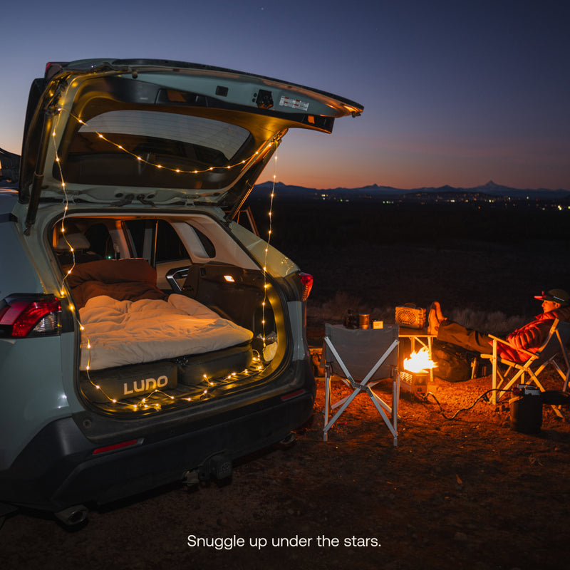 A car with its trunk open, decorated with string lights, showcases a cozy bed setup featuring the Luno® Fitted Sleeping Bag Comforter. Nearby, a person relaxes by a campfire in front of mountains under the starry sky, with camping chairs and a table. Text reads, “Snuggle up under the stars.”.