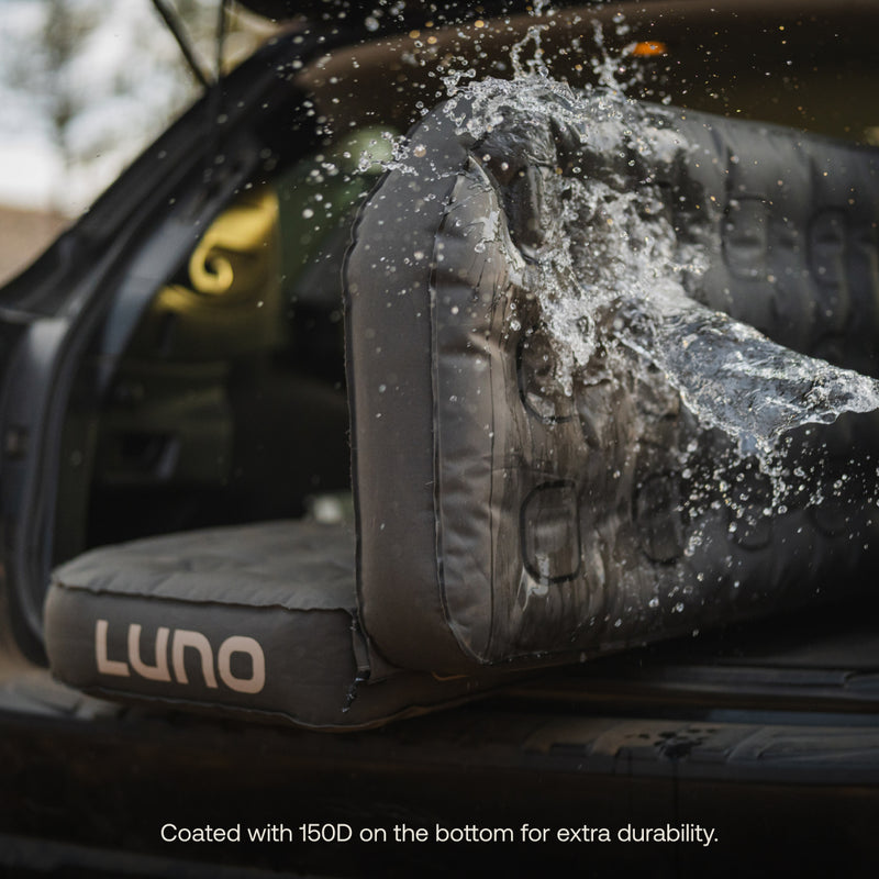 The Luno® AIR Pro Vehicle Mattress, inflated in the open trunk, demonstrates water resistance as droplets splash on its 150D-coated surface. Designed for durability, it includes Head Support Bridge Attachments and a Solo Zipper for comfort while traveling.
