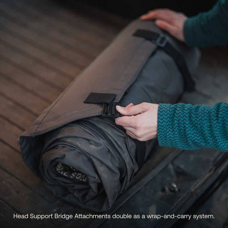 Someone in a teal sweater secures a strap on a rolled-up, gray sleeping bag that features "Head Support Bridge Attachments," doubling as a wrap-and-carry system, while the Solo Zipper by Luno® ensures the AIR Pro Vehicle Mattress stays intact.