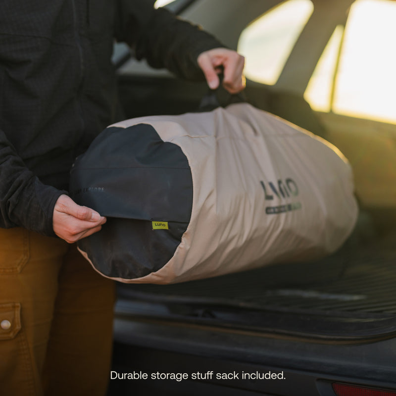 Someone carefully places a large rolled-up tarp in their vehicle, nestling it beside the Luno® AIR Pro Vehicle Mattress. The tarp is stored in a beige sack with a black handle. Text at the bottom says, "Durable storage stuff sack included.