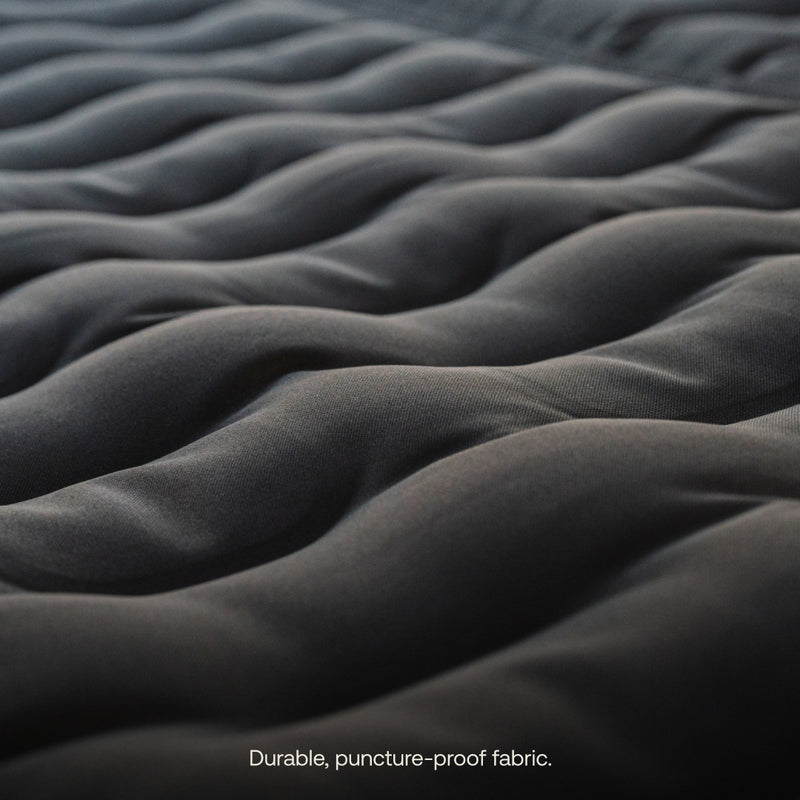 Close-up of the Luno® AIR Pro Vehicle Mattress shows a dark, wavy, quilted texture with a smooth, durable appearance. The puncture-proof fabric catches light and highlights contours.