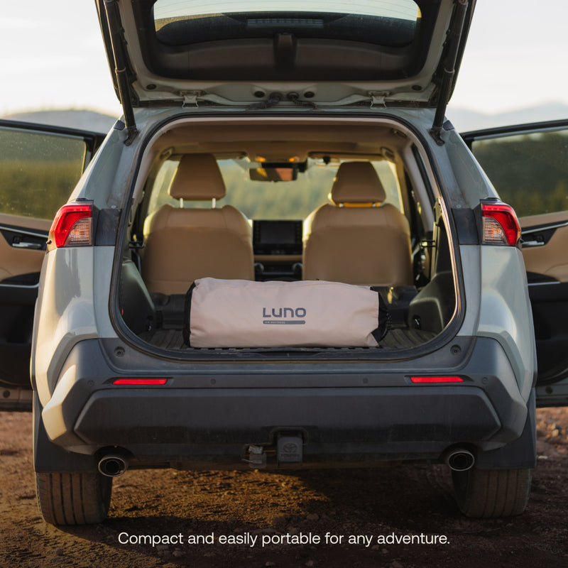 The image features the open trunk of a light gray SUV outdoors with a rolled-up Luno® AIR Pro Vehicle Mattress. Two tan seats are seen through the rear window. The caption states, "Compact with Solo Zipper, easy to pack and portable for adventures.