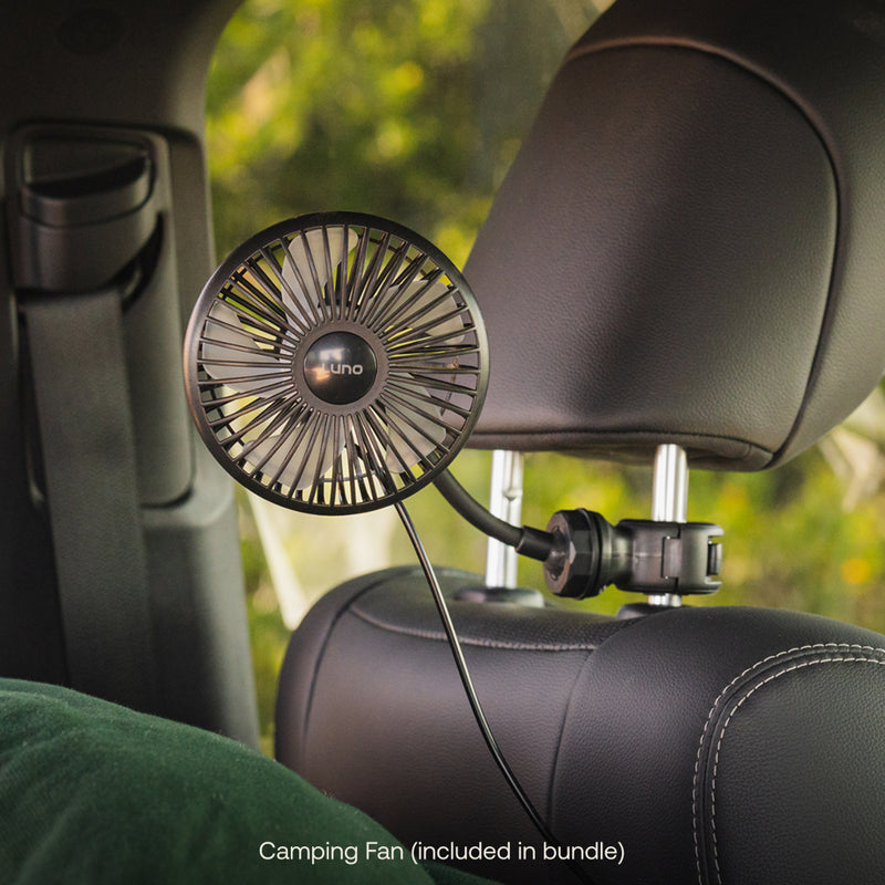 The Luno® Car Camping Cool Down Bundle, an essential for car camping, is clipped to a seat headrest facing the passenger area. Plugged in and ready to use, it helps keep you cool with a view of trees through the car window.