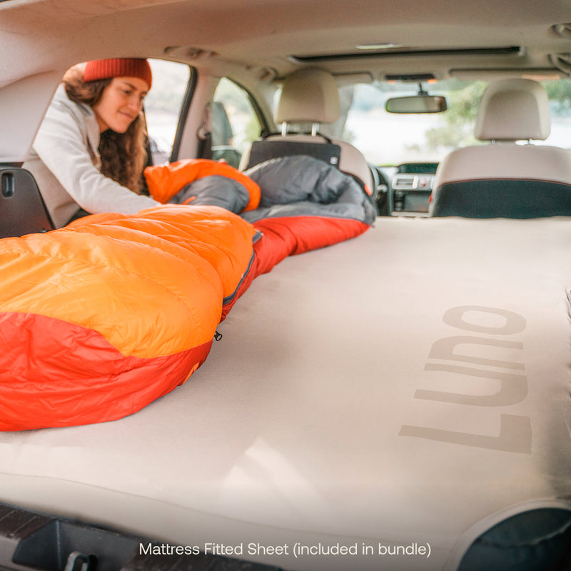 A woman arranges an orange sleeping bag on a Luno® Car Camping AIR Pro Starter Bundle in her car, getting ready for camping. Essentials like the included mattress fitted sheet boost comfort for vehicle adventures.