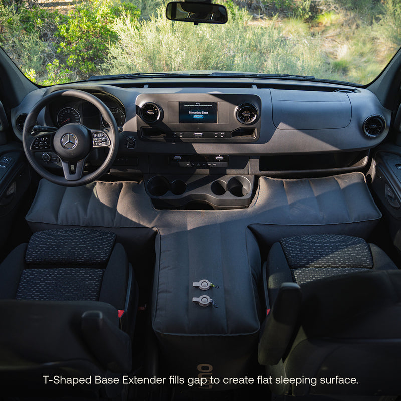 View of a van’s front seats with the Luno® AIR Pro Van Front Cab Mattress and padded T-shaped base extender creating extra sleeping space. Sunlight enters through the windshield, greenery outside. Easily set up using a cordless electric pump.