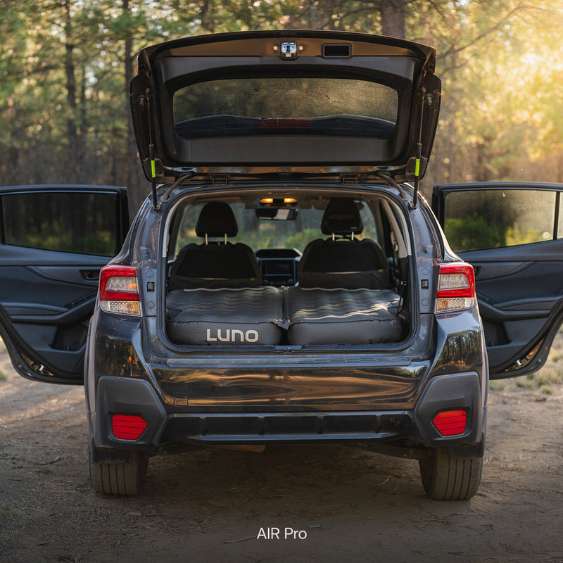 A grey SUV, the Subaru Crosstrek, is parked with its hatch and doors open. The back seats are folded down to fit the Subaru Crosstrek Car Camping Mattress, turning the cargo area into a comfortable sleeping space.