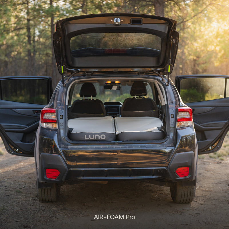 A Subaru Crosstrek with its trunk and rear doors open is parked in a sunlit forest clearing. The back seats are folded down, and a Subaru Crosstrek Car Camping Mattress is set up inside, ready for camping adventure.