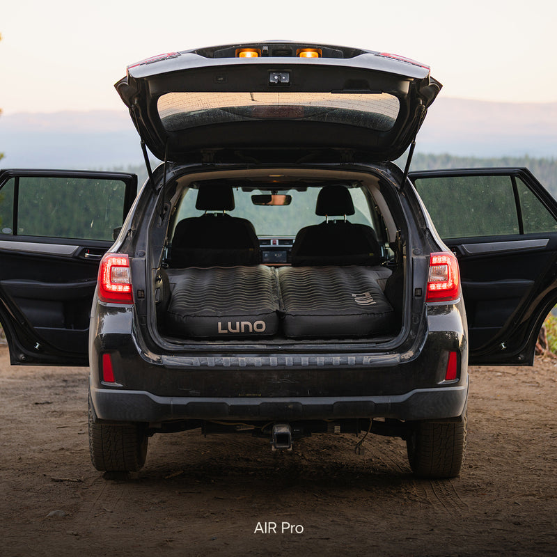 A Subaru Outback is parked outdoors with all doors open. The back seats are folded, and a Subaru Outback Car Camping Mattress is inflated in the rear cargo area, creating a comfortable sleeping space inside the vehicle.