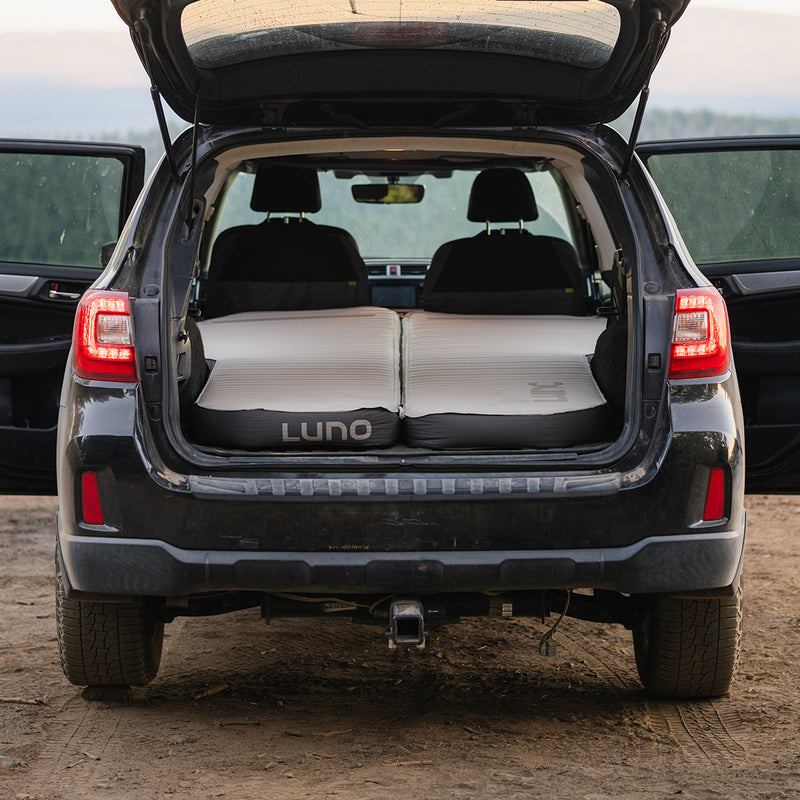 A black SUV with its rear hatch and side doors open displays a Subaru Outback Car Camping Mattress inflated in the back, seats folded down, perfect for car camping with a scenic outdoor view in the background.