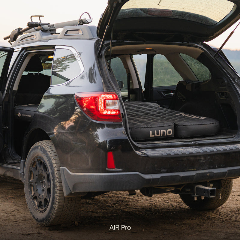 A black SUV with its rear hatch open displays a Subaru Outback Car Camping Mattress fully set up in the cargo area, ready for an outdoor adventure. The vehicle is parked on a dirt surface.