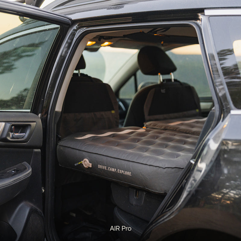 The Subaru Outback Car Camping Mattress is set up in the backseat of a black car, stretching from rear to front seats, with the label DRIVE. CAMP. EXPLORE. visible on the mattress.