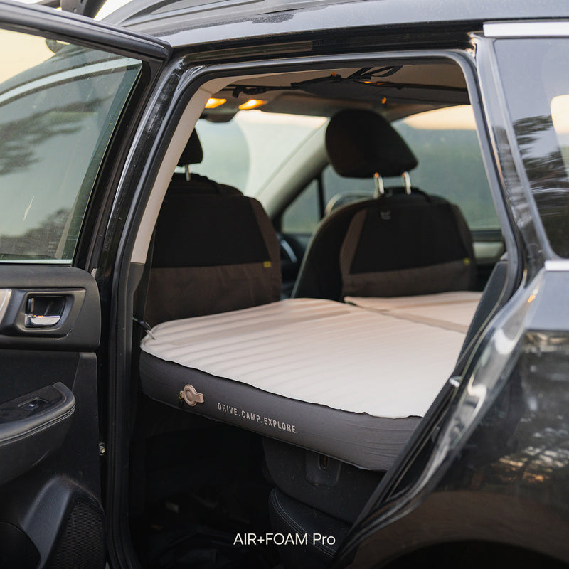 A Subaru Outback with its back door open shows a Subaru Outback Car Camping Mattress covering the back seat area, labeled DRIVE.CAMP.EXPLORE. and AIR+FOAM Pro—perfect for car camping.