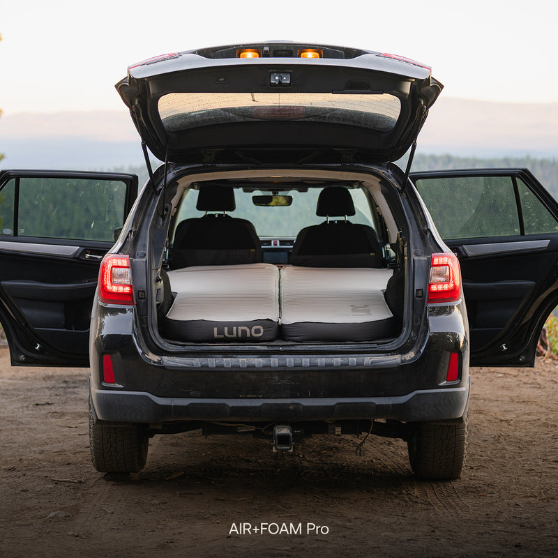 A black SUV with open rear doors and hatch showcases an inflated Subaru Outback Car Camping Mattress in the cargo area, with a natural outdoor background.
