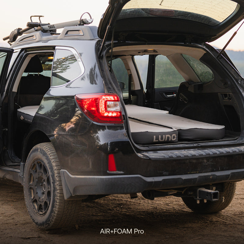 A black SUV with its tailgate open reveals a folded Subaru Outback Car Camping Mattress set up in the cargo area, ready for car camping. The vehicle is parked outdoors on a dirt surface.
