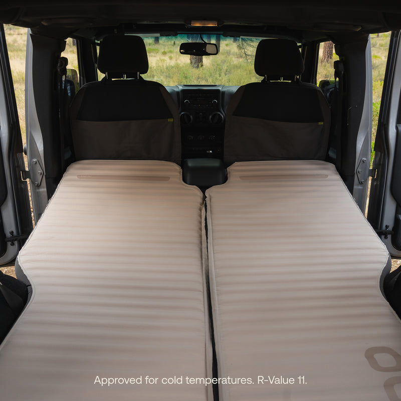 Two AIR+FOAM Pro Jeep Wrangler Mattresses create a cozy sleep setup in a vehicle’s back. Front seats and open doors show an outdoor scene. Text at bottom: Approved for cold temps. R-Value 11. Perfect Jeep Wrangler mattress solution.