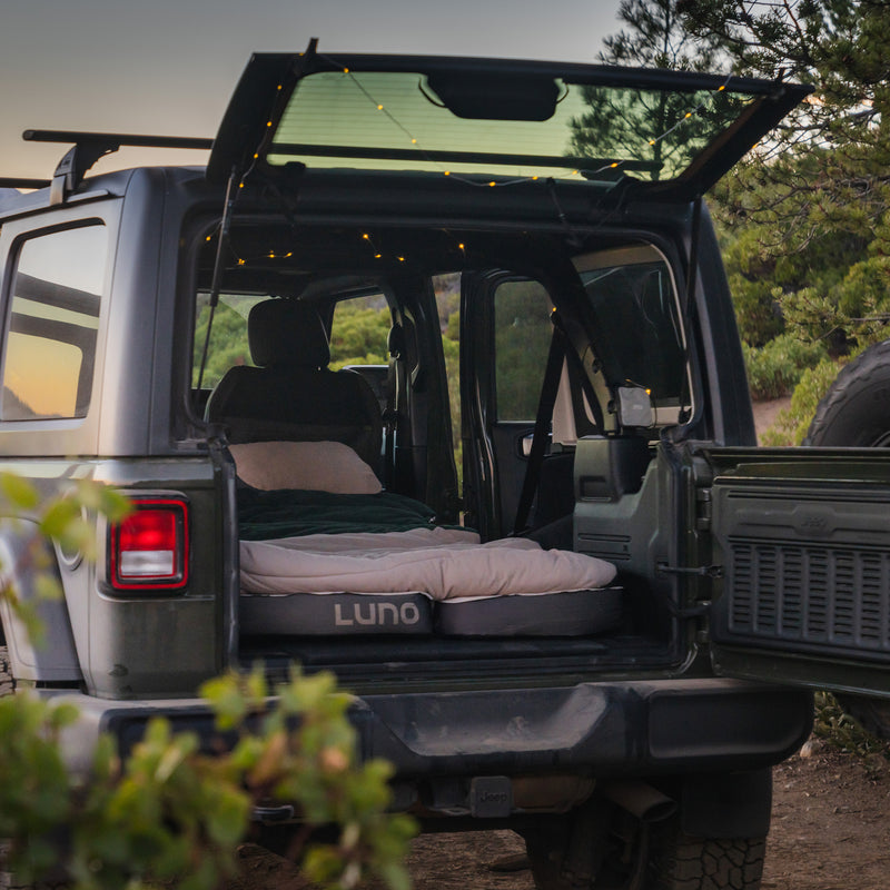 A Jeep Wrangler with its rear door open displays a comfy AIR+FOAM Pro Jeep Wrangler Mattress and string lights inside, set outdoors among greenery at sunset.