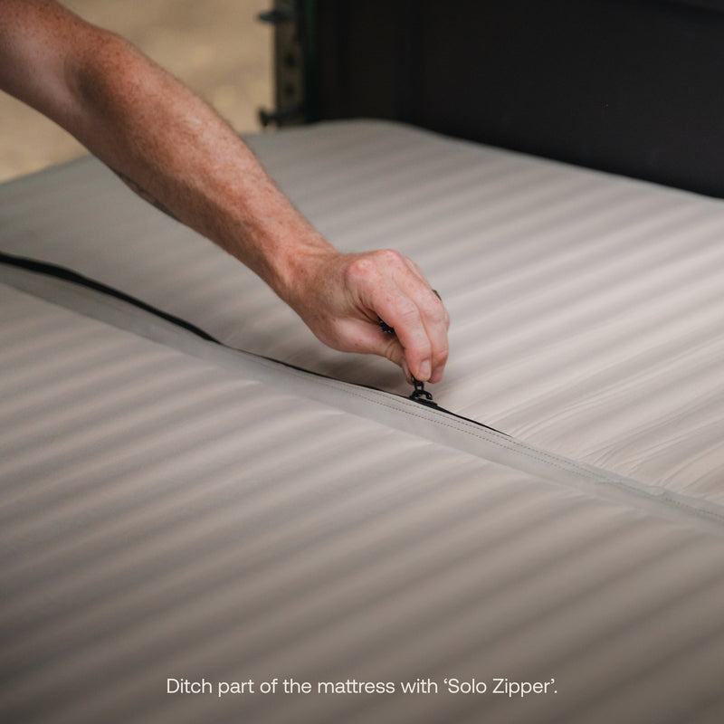 A close-up of a hand unzipping the AIR+FOAM Pro - Rivian R1S Compatible mattress seam, demonstrating the “Solo Zipper” feature. Text at the bottom: “Ditch part of the mattress with ‘Solo Zipper’.”.