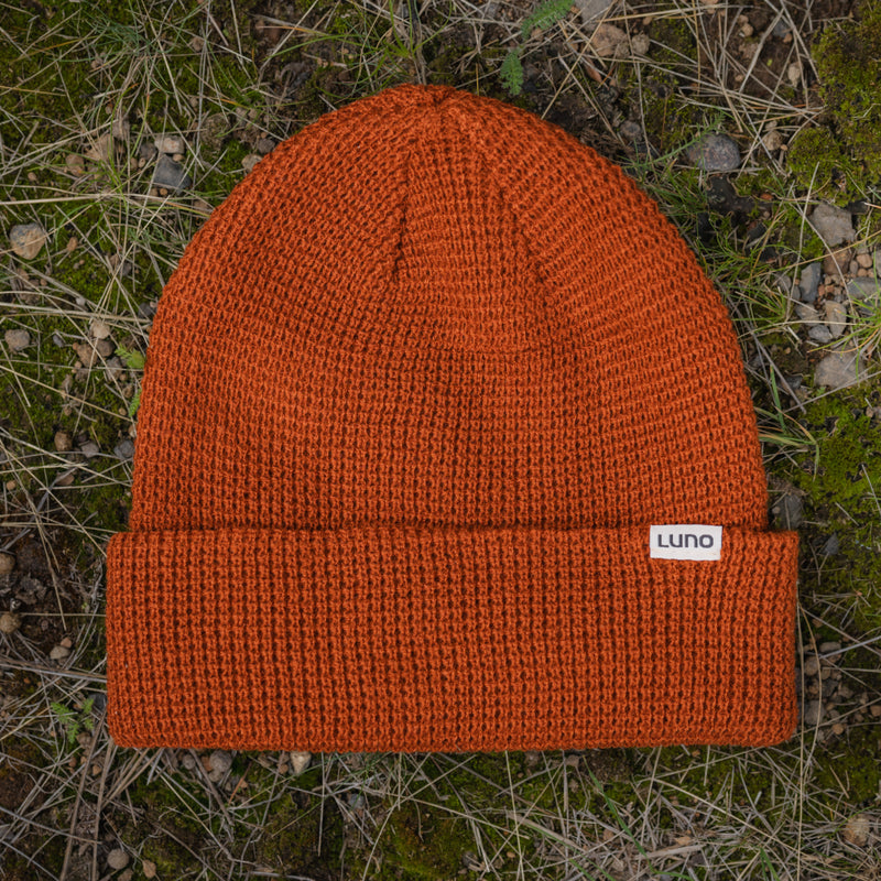 The Waffle Knit Camp Beanie, featuring an orange waffle knit design, a folded brim, and a small white Luno tag, rests on dry grass and green moss.