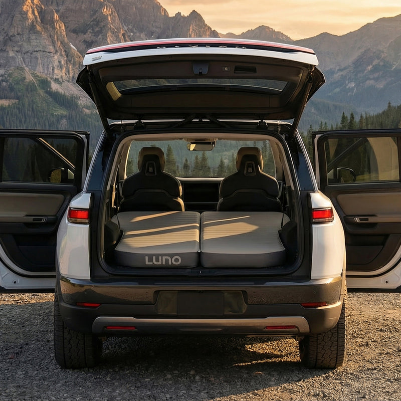 A white SUV with its rear hatch and side doors open is parked by mountains. The back seats are folded down and covered with a Luno AIR+FOAM Pro - Rivian R2 Compatible mattress, creating a flat sleeping space.