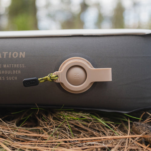 Close-up side view of an inflatable camping mattress on the ground. The focus is on the air valve, which is closed and has "IN" marked on it. The mattress rests on pine needles and grass, indicating an outdoor setting.