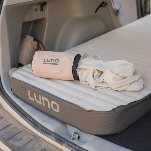 The image shows the back of an SUV with its trunk open. Inside, a Luno air mattress is fully inflated, and a partially unrolled Luno storage bag with bedding lies on top of it. The setup looks ready for car camping or sleeping in the vehicle.
