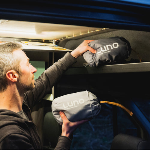 A person in a van is storing two gray bags labeled "Luno" on an overhead shelf. One bag is already placed on the shelf, and the person is lifting the second bag to store it next to the first. The van interior has a cozy feel with string lights.