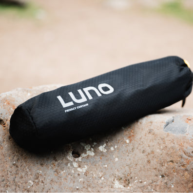 A black cylindrical bag with the word "Luno" and "Privacy Curtain" printed on it rests on a rocky surface outdoors. The background is slightly blurred, showing a mix of dirt and vegetation.
