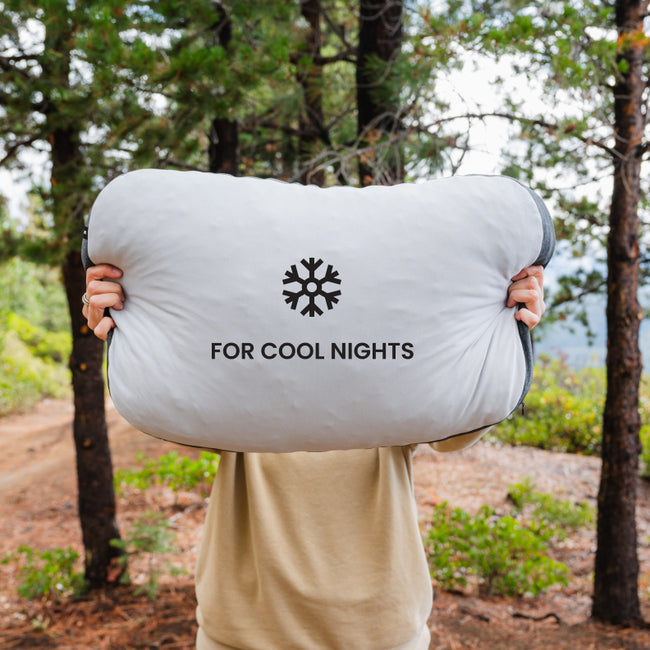 Person standing in a wooded area holds a large pillow in front of their face. The pillow is light gray with a black snowflake symbol and the text "FOR COOL NIGHTS" printed on it. The background features trees and a dirt path.