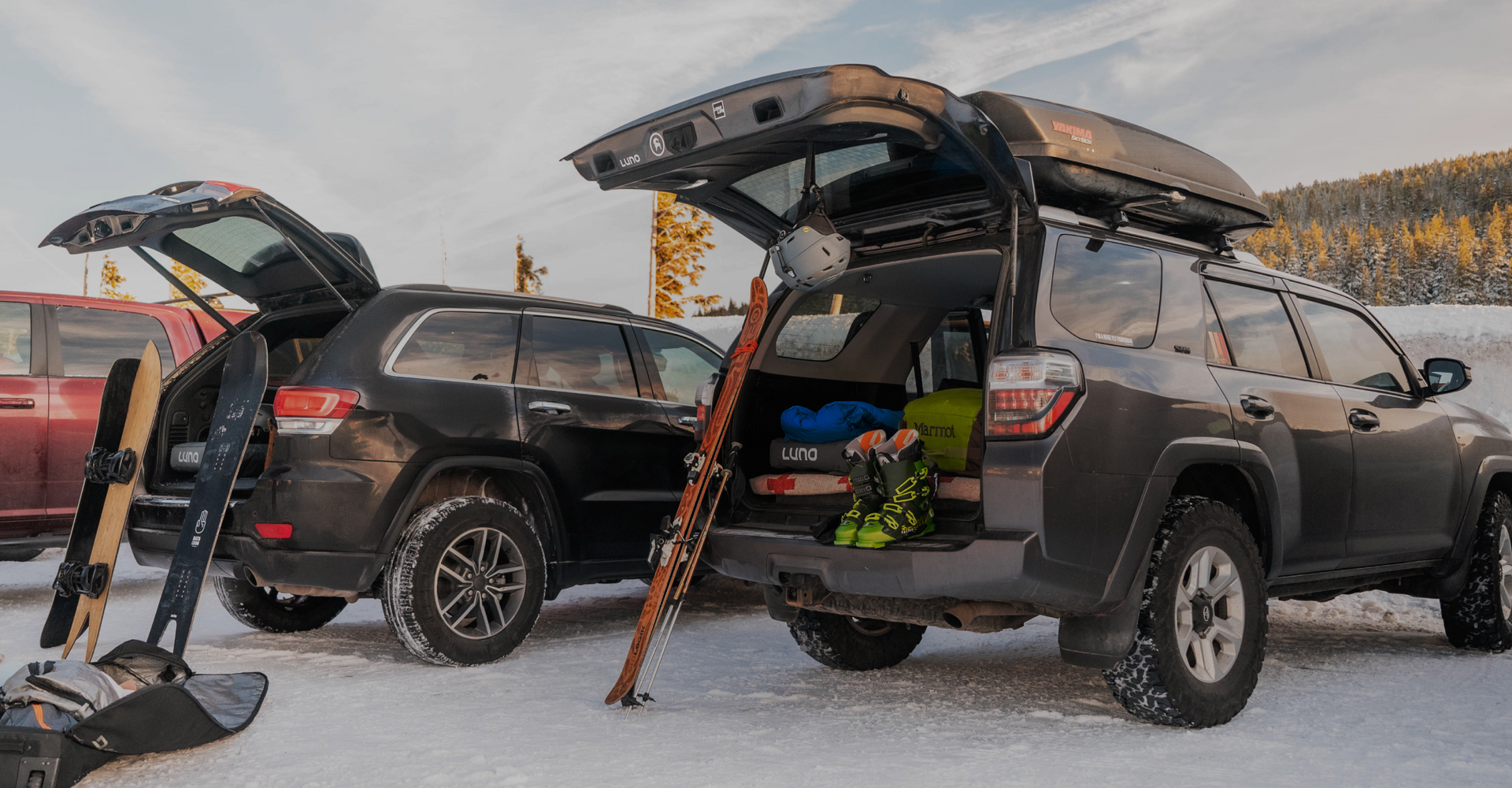 Two SUVs are parked in snowy conditions with their rear doors open. Skis, snowboards, and winter gear are visible in the back of the vehicles. The sun is setting, casting a warm glow on the trees in the background. A third vehicle is partially visible to the left.