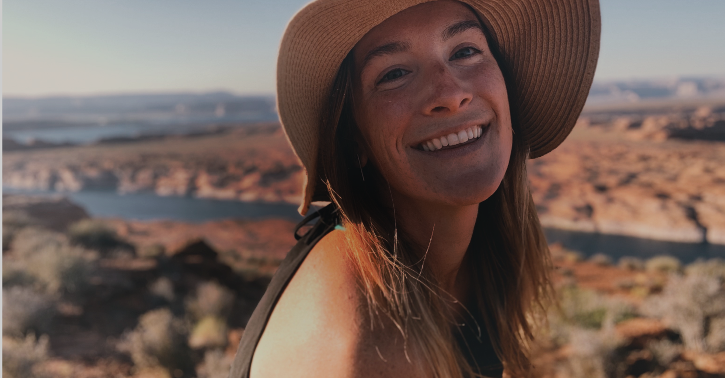 A woman with long brown hair and a wide-brimmed hat smiles at the camera. She is outdoors, with a scenic background of a rocky desert landscape and a river in the distance under a clear blue sky.