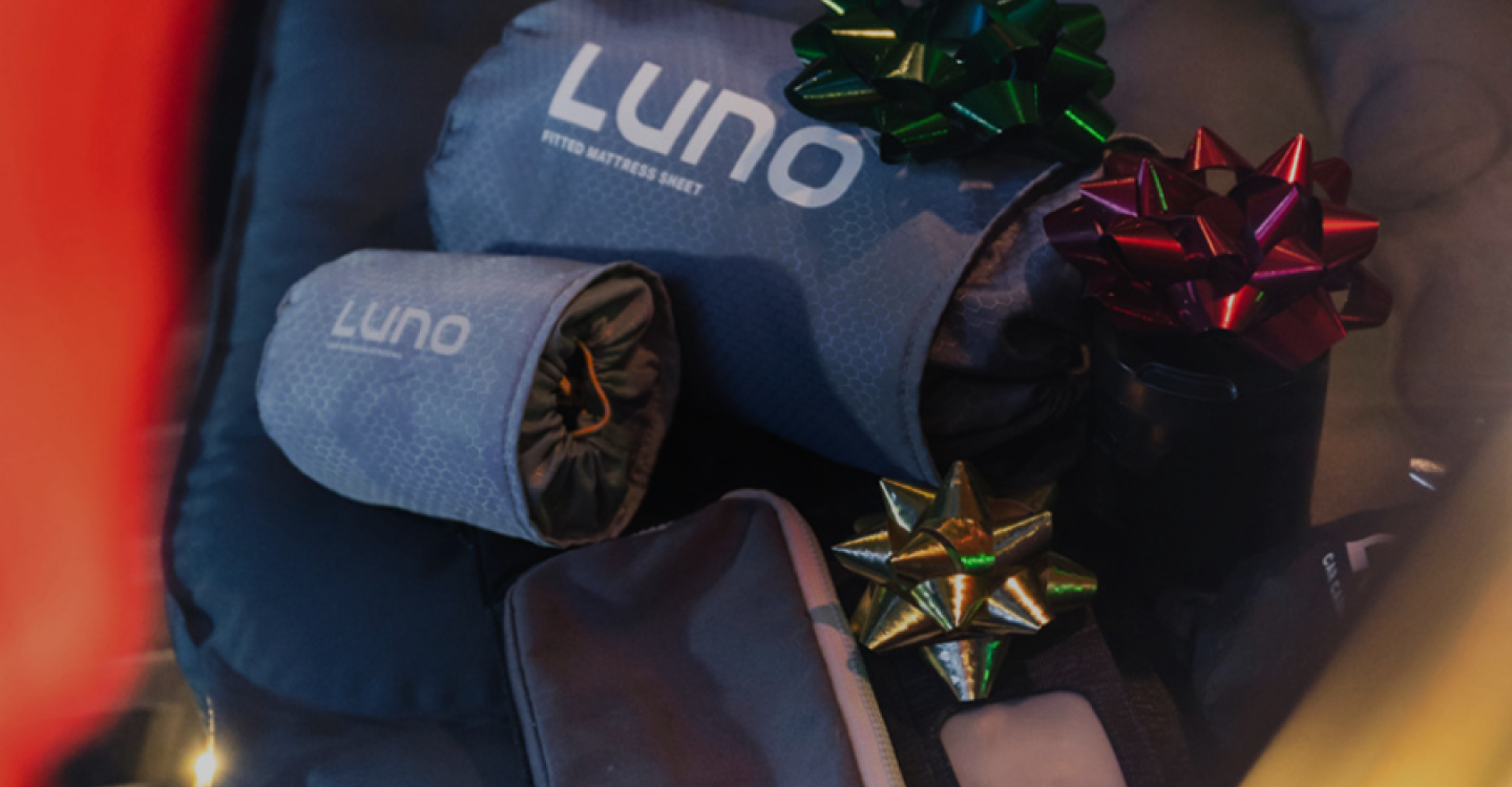 A display of Luno brand camping gear, including a fitted mattress sheet and other items, each adorned with colorful gift bows. The equipment appears neatly arranged, suggesting a holiday or gift-giving theme.