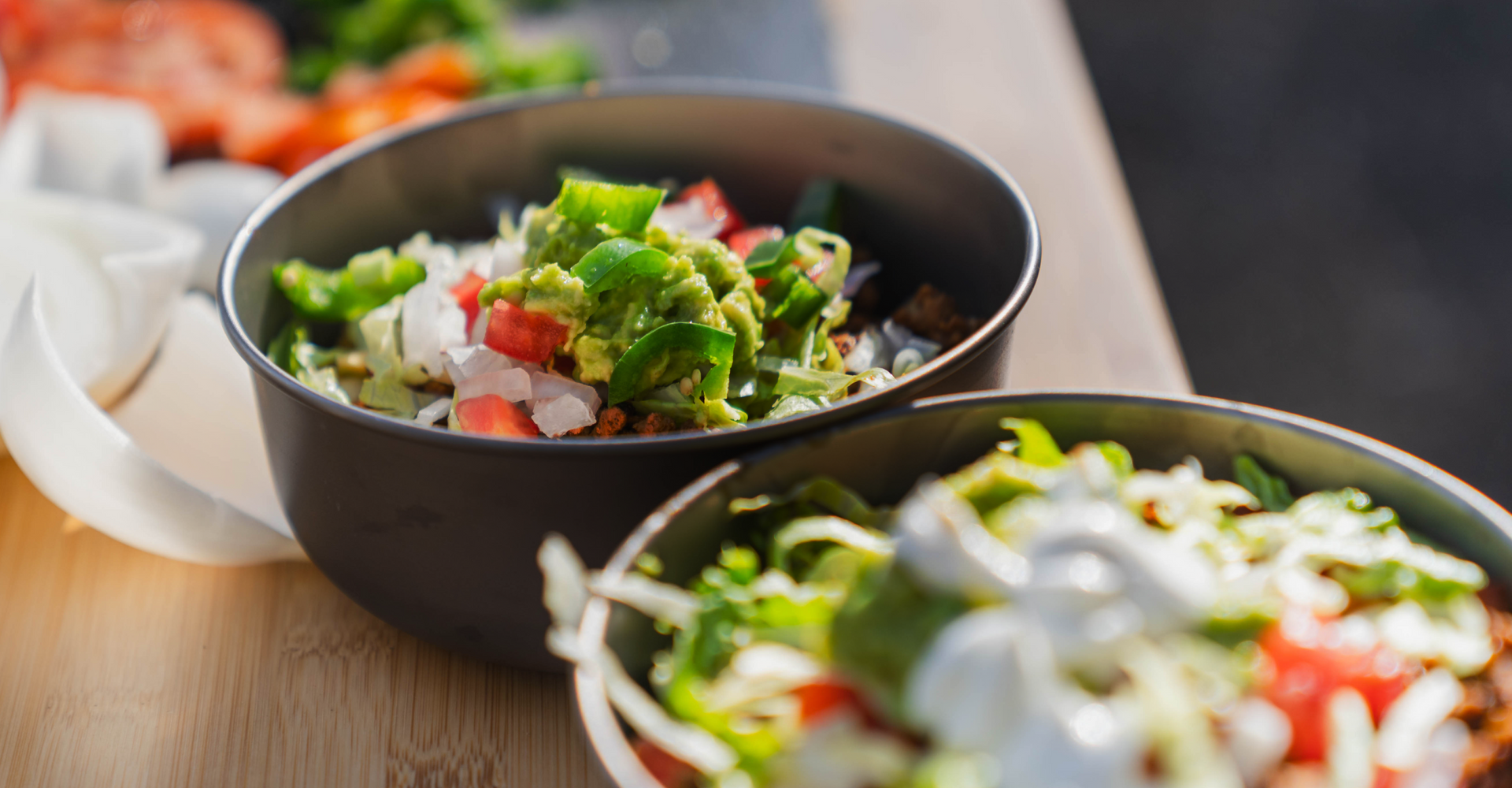 Two bowls of fresh salad are placed on a wooden surface. Each bowl contains a colorful mix of vegetables, including tomatoes, lettuce, onions, and what appears to be guacamole and a dollop of sour cream or a similar topping. More chopped vegetables are in the blurred background.