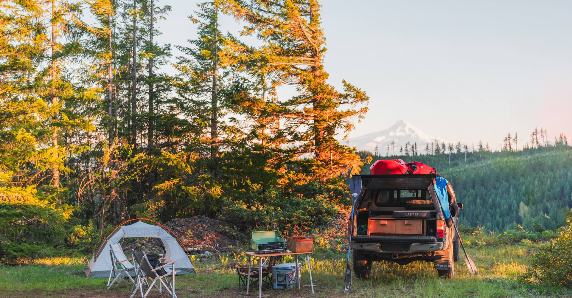 A scenic camping setup includes a pitched tent, folding chairs, and a table with supplies in a forest clearing. A truck with gear is parked nearby, with the hood facing a backdrop of mountains and trees. The sky is clear, suggesting early morning or evening light.