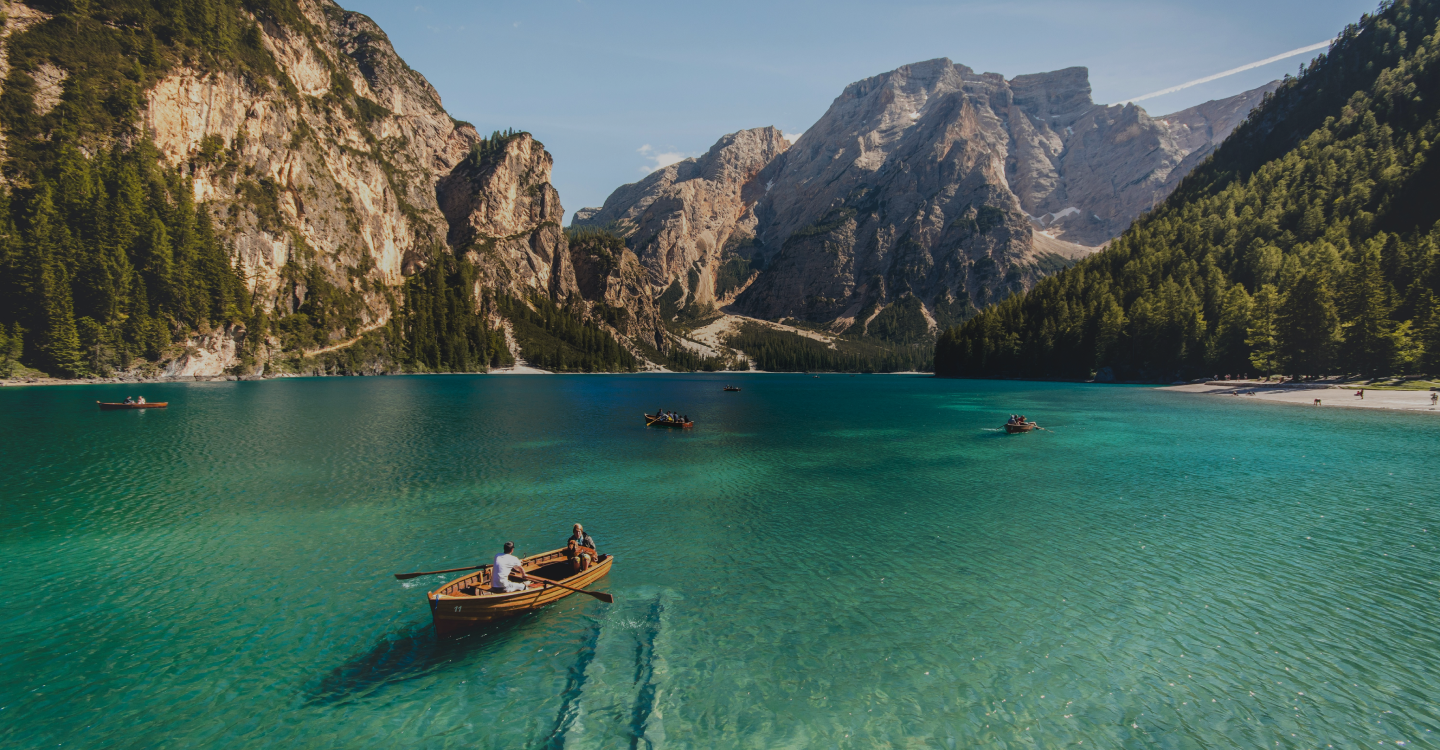 A serene alpine lake surrounded by towering mountains, with clear turquoise water reflecting the blue sky. Multiple wooden rowboats, each with people inside, are scattered across the lake. Dense green forests cover the slopes, leading up to rocky cliffs.