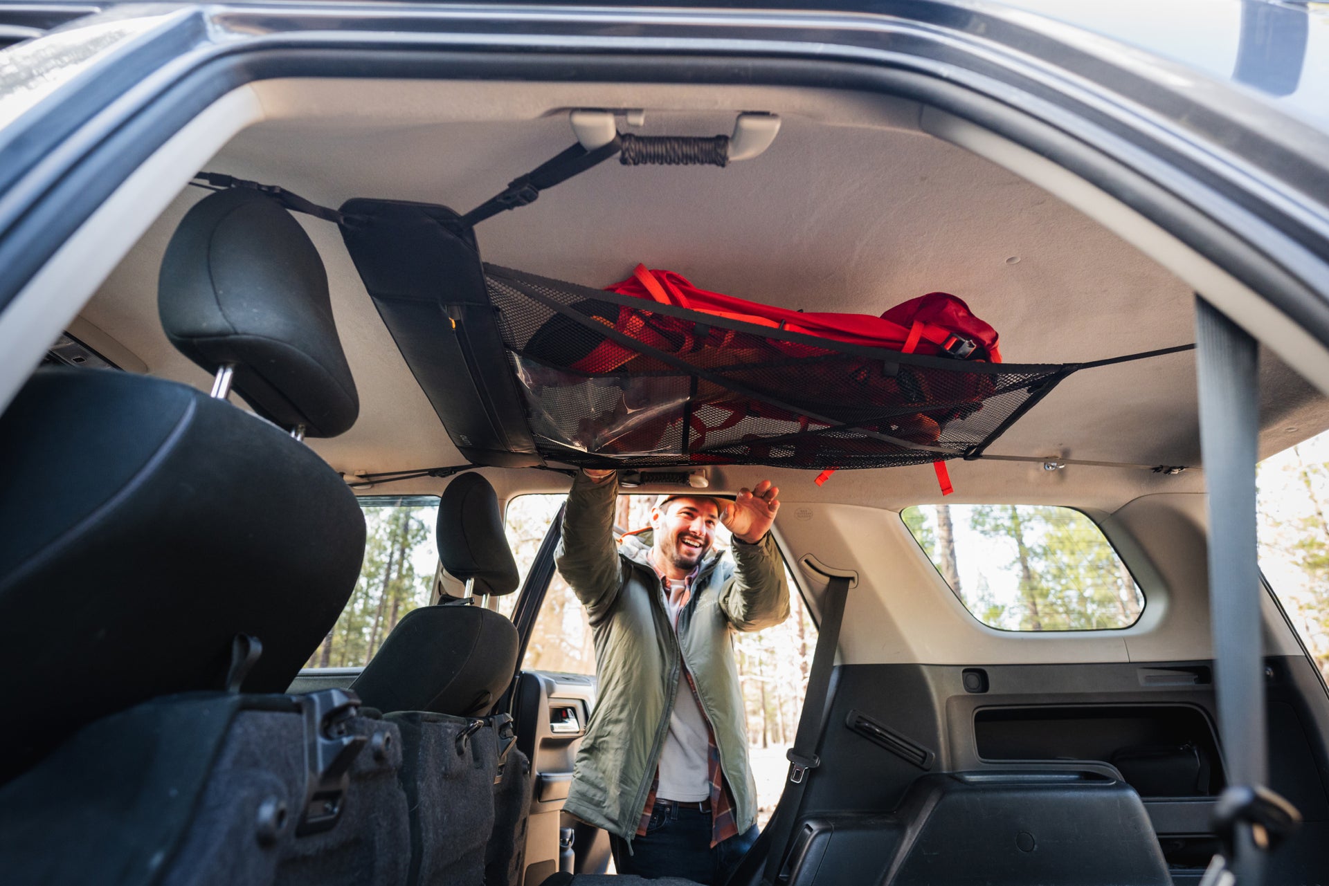 A person standing outside an open car door, reaching up to place or retrieve a red backpack from a netted storage compartment on the interior roof of the car. The car's interior is spacious, and sunlight filters through the windows, highlighting the organized storage area.