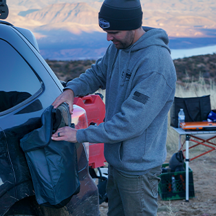 A man in a grey hoodie and beanie sets up camp next to his car in an outdoor setting with mountains and a lake in the background. He is attaching a bag to the vehicle. Camping gear is visible on a table nearby.