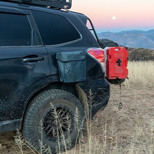 A black SUV with off-road tires is parked in a dry, grassy area near hills and mountains. The vehicle has a red fuel canister and a black storage bag attached to the rear. A full moon can be seen in the twilight sky.