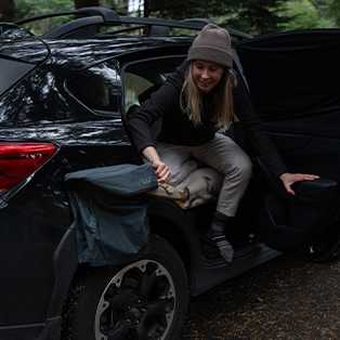 A person dressed in warm clothing, including a beanie and socks, sits partially inside a dark vehicle with an open back door. The person appears to be getting comfortable, possibly preparing for a rest or overnight stay in the car. A forest setting is in the background.