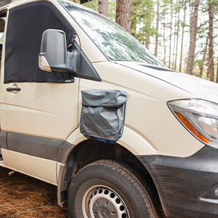 A beige camper van is parked in a wooded area. A gray storage bag is attached to the outside of the driver's door. The side mirror, window cover, and part of the van's wheel are visible. Tall trees are in the background.