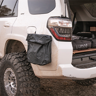 A white SUV with mud on its body and a large off-road tire is parked, the rear hatch open. A black storage bag labeled "Luno" is attached to the back of the vehicle. The interior reveals more camping gear, also labeled "Luno.