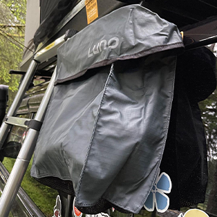 A gray Luno storage bag is attached to a metal ladder on a camper. The background features trees and outdoor scenery, suggesting the setting is a campsite. The bag appears to be made of durable, weather-resistant material.