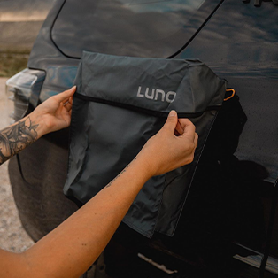 A person with tattooed arms attaches a black Luno storage bag to the back of a black car. The bag is rectangular with a flap and appears to be made of a water-resistant material.