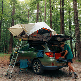 A person stands at the back of a green SUV with an open trunk in a forest. The car is equipped with a roof-top tent, which is open and accessible via a ladder. Various camping gear and a folding chair are visible around the vehicle.
