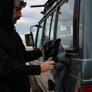 A person in a black hoodie and beanie packs gear into a gray bag beside a rugged off-road vehicle on a cloudy day. The vehicle appears to be on an outdoor adventure, set against a backdrop of sandy terrain and a cloudy sky.