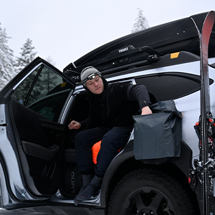 A person dressed in winter outdoor gear is sitting in the backseat of a car with the door open, holding a gray bag. The car has skis attached to the side and a roof rack. The background features snow-covered trees.